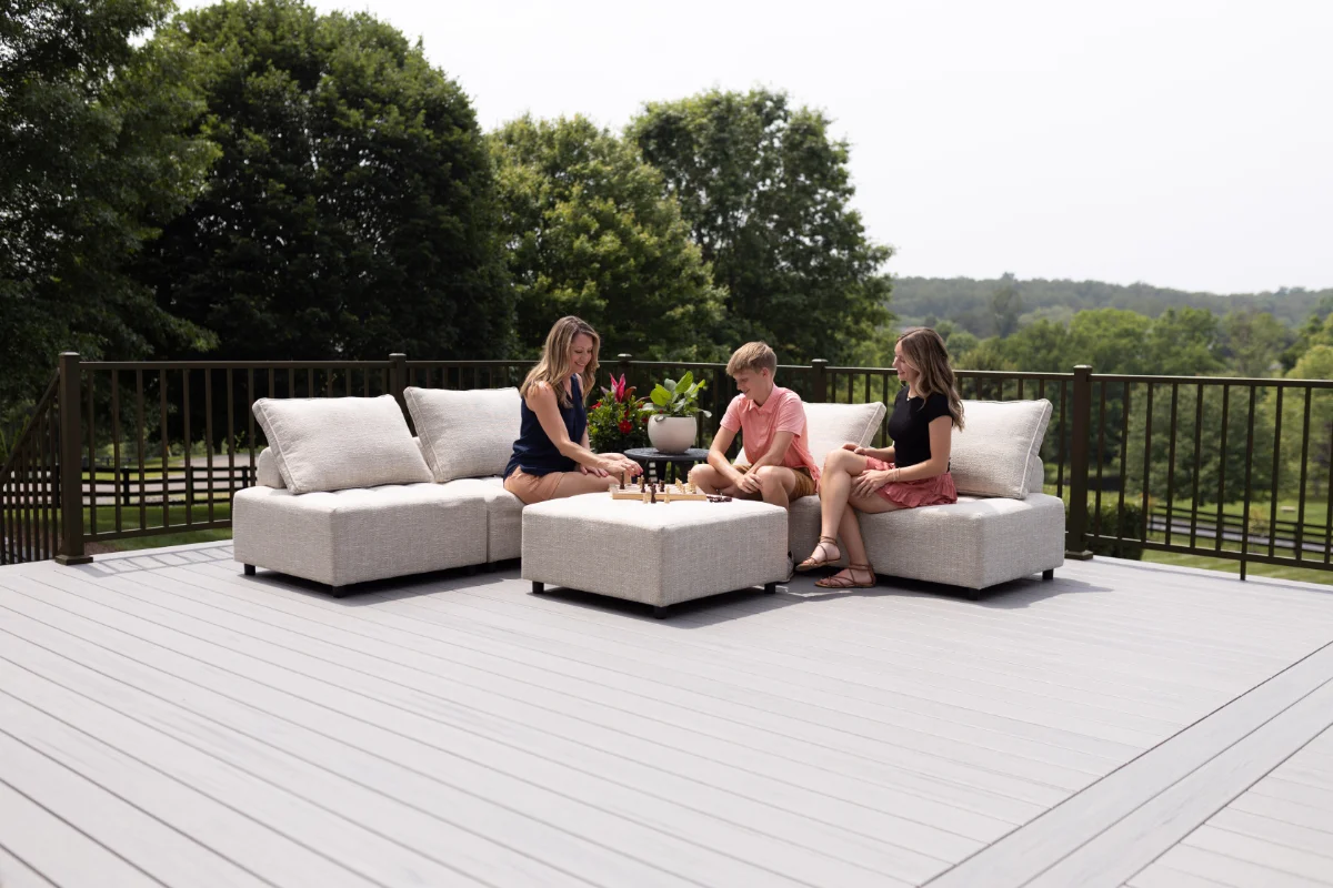 Three people—an adult woman, a child, and another adult woman—sit on a beige outdoor sectional on a wooden deck, playing chess with a potted plant centerpiece and a green landscape in the background.