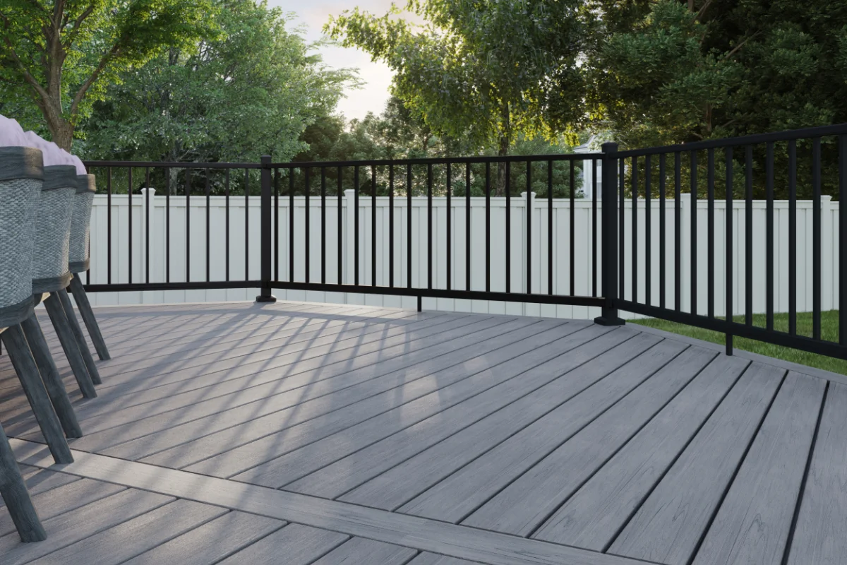 Trex-decking-img-2 Wooden outdoor deck with a black metal railing and a row of dining chairs on the left, white privacy fence in the background, and trees beyond.