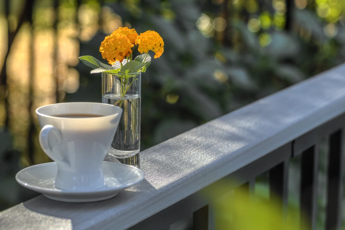 Trex-railing-img-3 White coffee cup on a saucer beside a glass vase with yellow flowers on a sunlit railing
